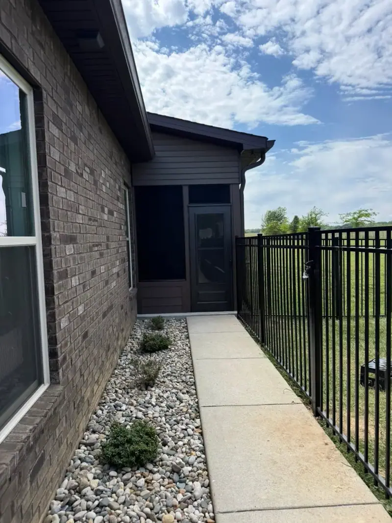 Concrete walkway beside a brick home with river rock and black fencing