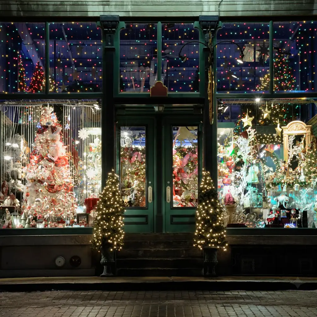Storefront lit for the holidays at night, with color lights across the transom windows, decorated window displays and lit trees at the door