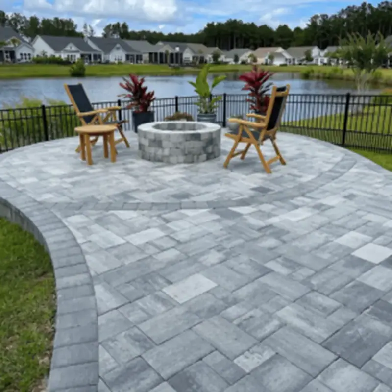 Curved paver patio with a stone fire pit and chairs overlooking a pond